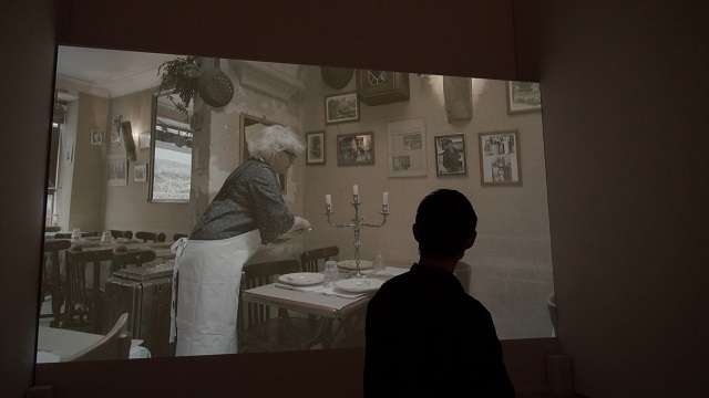 映像作品《着る喜びの風景》の一コマ。解説する皆川さんと。作品の舞台はフランスにある行きつけのビストロ